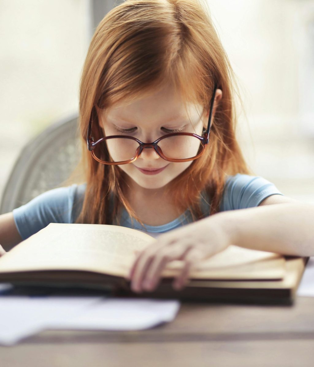 Kleines Mädchen mit roten Haaren und einer großen Brille liest ein Buch